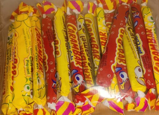 Carambar Caramel, Cola & Citron Piquant – Bonbons Tenders Français Bonbons colorés emballés, principalement jaunes et rouges, avec des motifs variés.